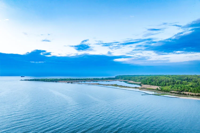 Aerial view of the Chesapeake Bay shoreline in Maryland, showing wooded coastal land, sheltered water, and narrow breakwaters beneath a cloudy sky.