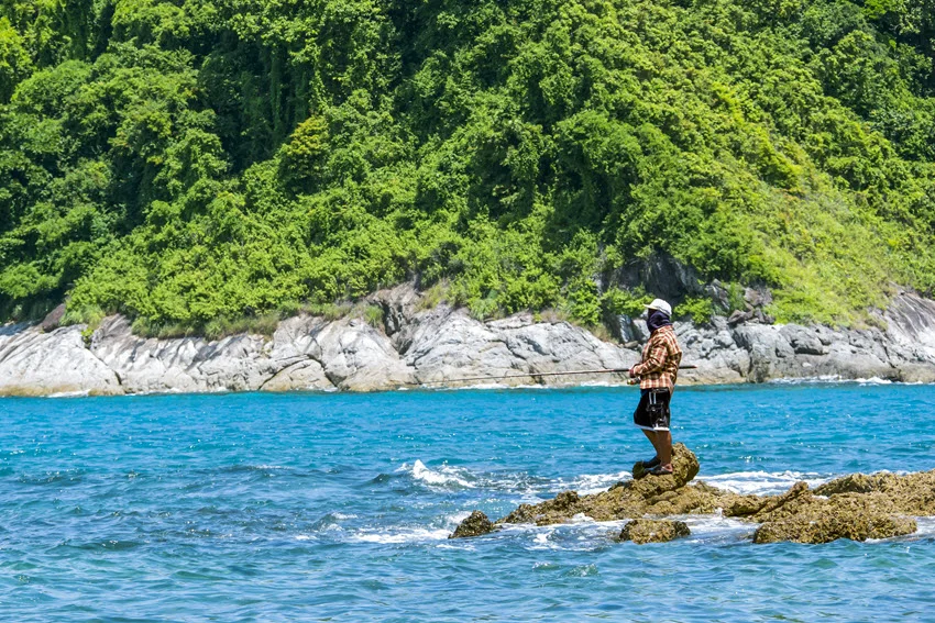 Angler holding a rod from coastal rocks above turquoise water, illustrating a shore-fishing setting for jigging beside a densely wooded coastline.