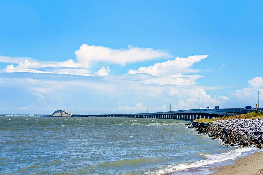 Dauphin Island Bridge in Alabama stretches across coastal water, with its raised span in the distance and a rocky shoreline in the foreground.