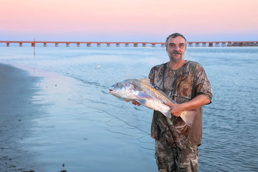 Angler holding a Black Drum beside a bay at sunset, with a long bridge and pink evening sky in the background.
