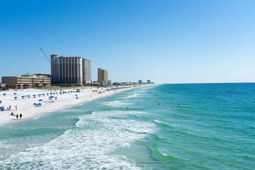 White sand and turquoise surf along Pensacola Beach on Florida's northern Gulf Coast, with beachfront buildings, umbrellas, and people near the water.