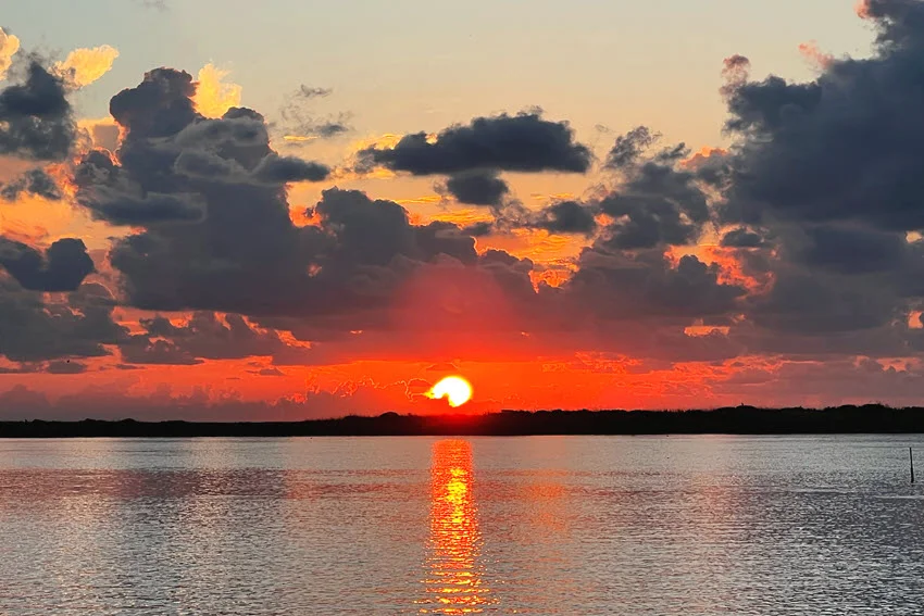 Orange sunrise over the Texas Gulf Coast, with dark clouds above a low shoreline and sunlight reflecting across calm coastal water.