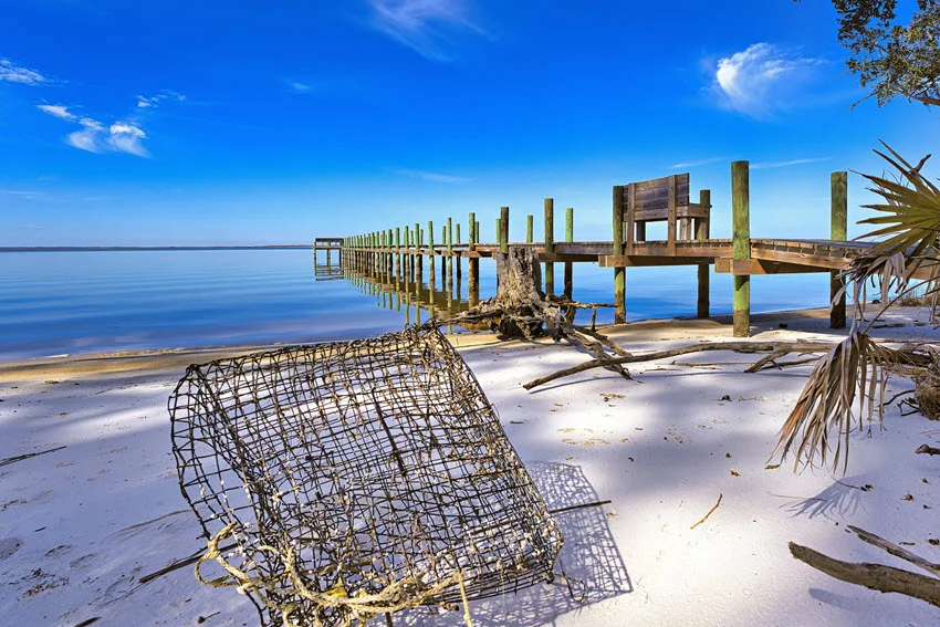 Wide coastal view of a quiet sandy shoreline on Apalachicola Bay, Florida, with a weathered fishing dock and a crab trap on the beach.