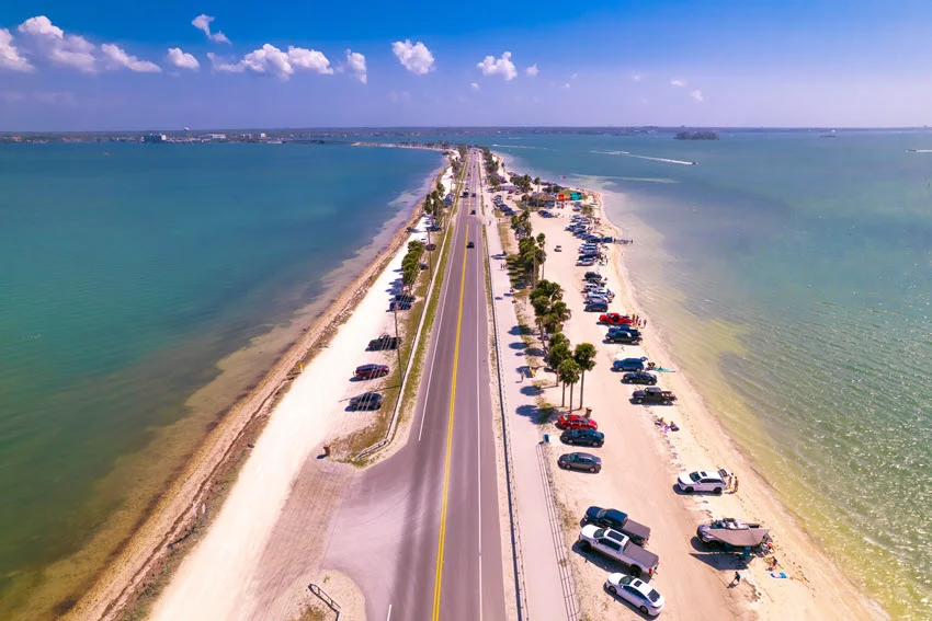 Aerial view of Honeymoon Island and the Dunedin Causeway in Florida, showing clear Gulf water, sandy shoreline, and beach access along the road.
