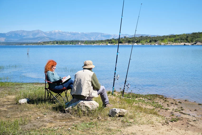 A man and a woman sitting by a calm lake; the man watches his fishing rod while the woman reads a book, creating a peaceful and relaxing scene.