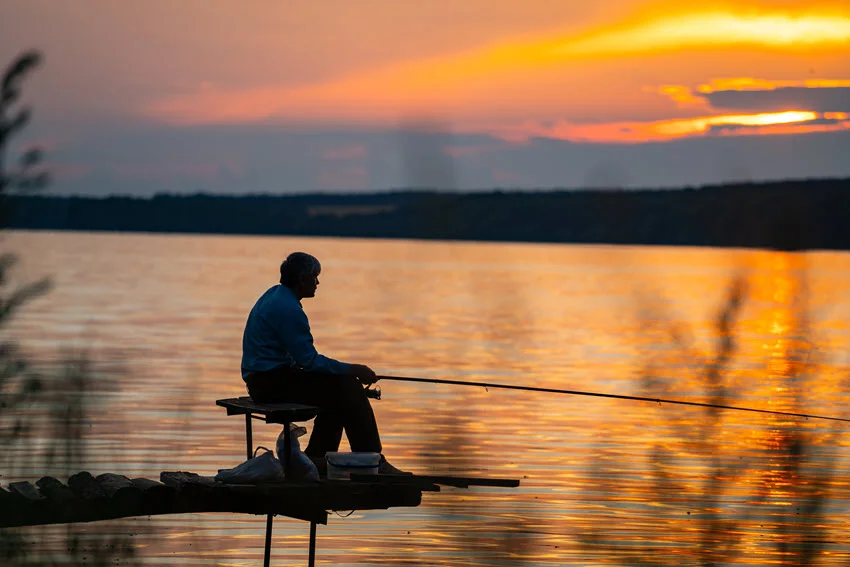 Silhouette of a fisherman casting a line from a dock at sunset, with a vibrant orange and yellow sky reflected on calm water.