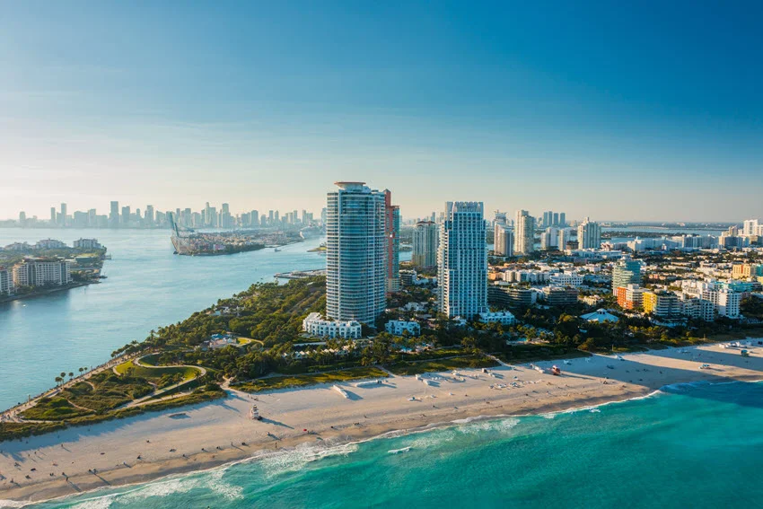 Wide drone shot of the Port of Miami, Florida, on a bright sunny day, showing panoramic aerial view of Miami Beach, South Beach, river, buildings, and lush vegetation.