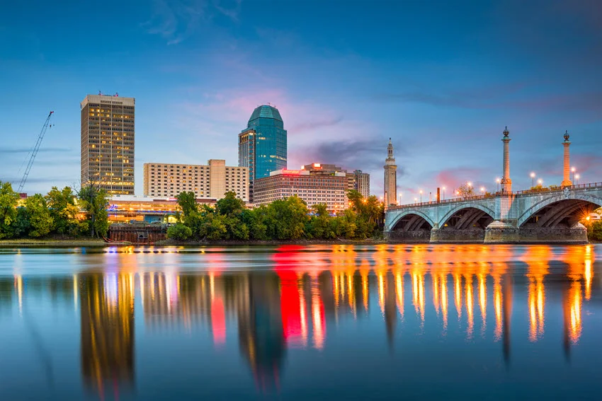 Springfield, Massachusetts, USA downtown skyline reflecting on the river at dusk, with city lights and a calm evening atmosphere.