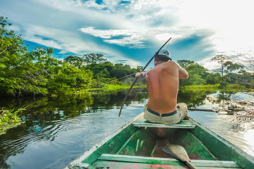 A young angler is sitting on a river-floating boat, aiming a bow and arrow at a fish surrounded by beautiful greenery and tranquility.