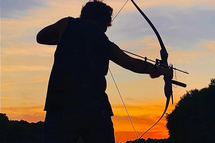 Close-up silhouette of an angler using bowfishing gear on a lake at night, with the sunrise casting a warm glow in the background.