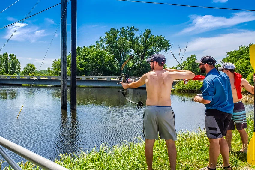 The angler is standing on a river shore with two friends, preparing to perform bow fishing.