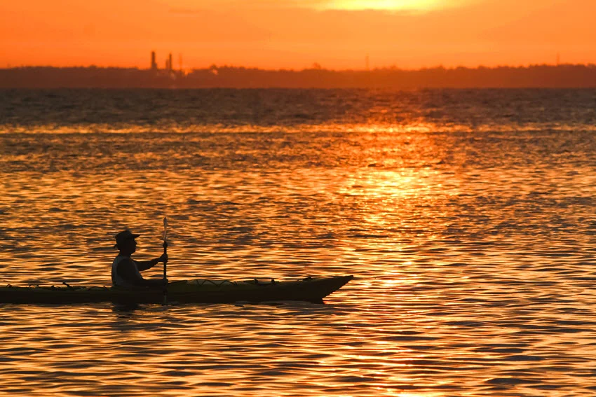 Silhouette view of an angler in a kayak gliding across the ocean during sunset.