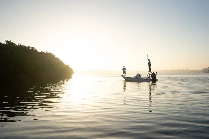 Wide view of two anglers fishing from a shallow water skiff at sunrise near mangroves in calm coastal water.