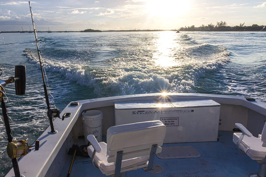 View from the stern of a fishing charter boat running across calm coastal water at sunset with rods set in holders and shoreline in the distance.