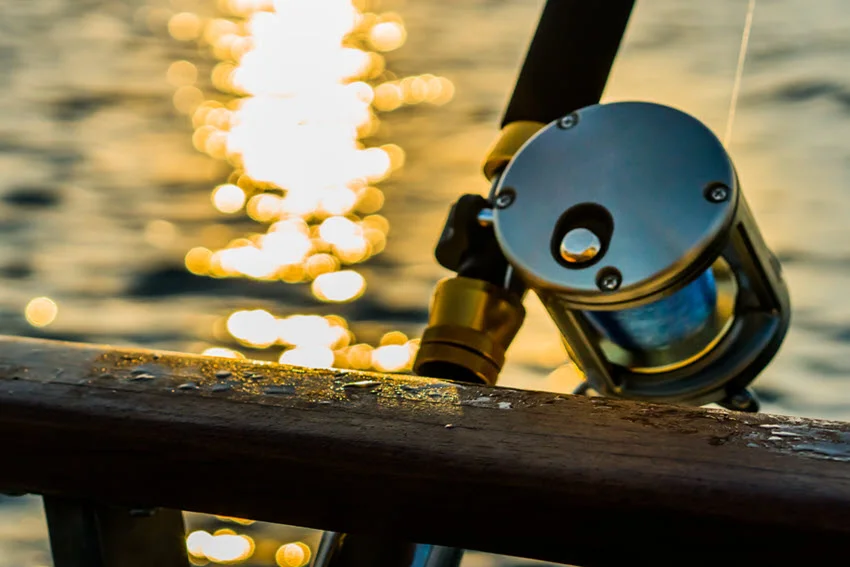 Close view of a fishing reel mounted on a boat rail at sunset with golden reflections on the water in the background.