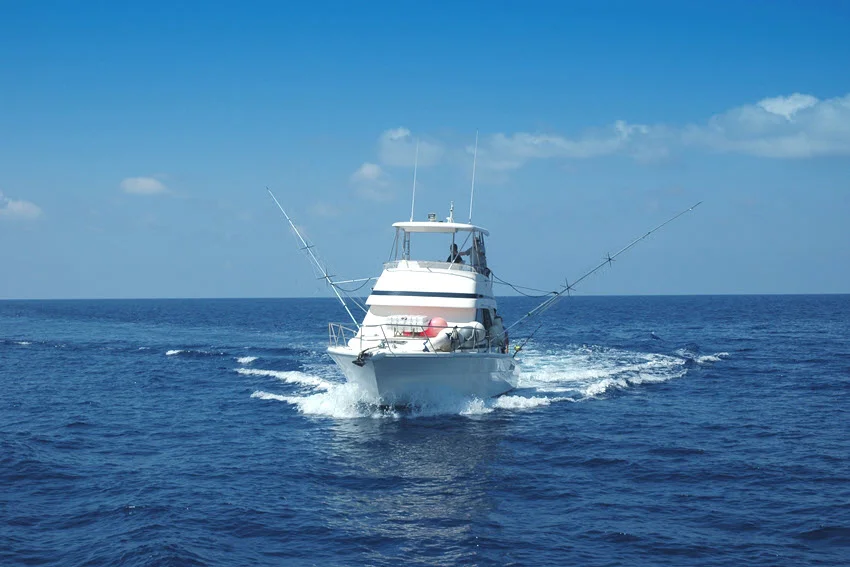 View of an offshore fishing charter boat running across open blue water with outriggers extended under a clear sky.