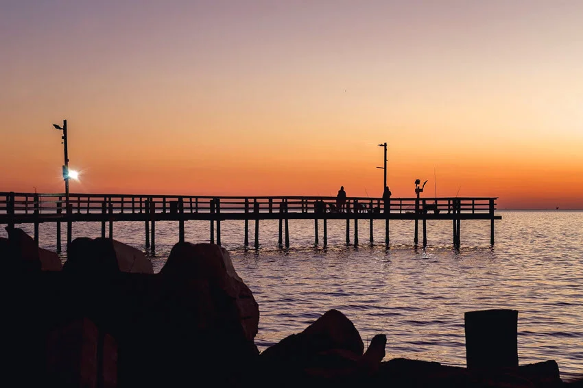 View of Cedar Point Pier in Bay St. Louis, Mississippi, at sunset with anglers fishing along the wooden pier over calm golden waters.