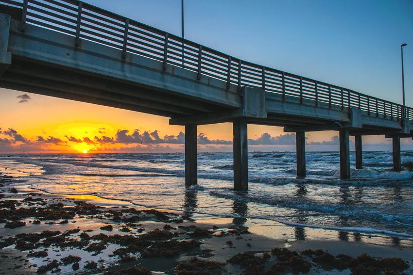 Pier along Padre Island in Corpus Christi, Texas, stretching over the Gulf of Mexico. The wooden structure offers a scenic spot for fishing and sightseeing, with waves gently rolling beneath and a vast coastal horizon in the background.