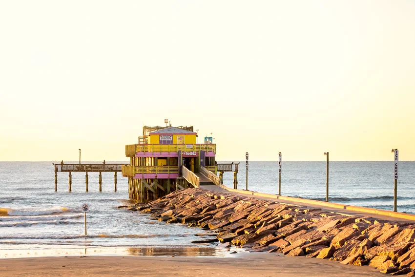 Sunrise at the 61st Street Pier on Seawall Blvd in Galveston, Texas. The early morning light casts a golden glow over the pier and ocean, creating a peaceful and picturesque coastal scene.