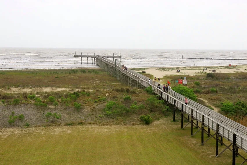 Louisiana nature background with a wooden boardwalk winding its way through sand dunes towards the beach and pier. The boardwalk offers a scenic path over the soft, golden sands, framed by coastal grasses, with the serene Gulf waters in the distance.
