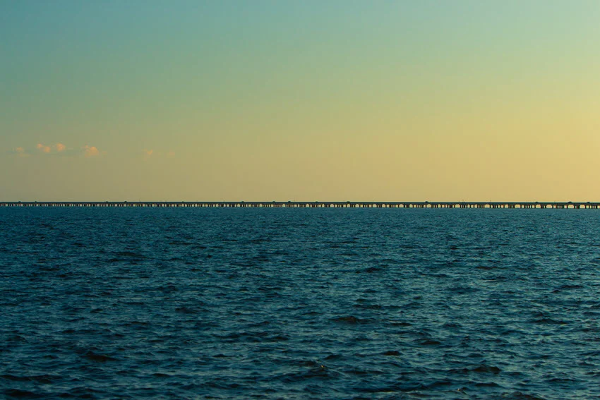 Sunset shot of the Lake Pontchartrain Causeway, one of the longest piers in the world. The golden and orange hues reflect off the calm water, while the endless stretch of the causeway fades into the horizon, creating a breathtaking and serene view.