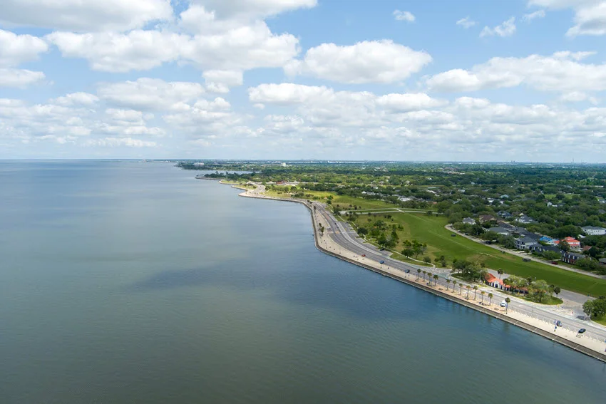 Aerial shot along the coast of Lake Pontchartrain in New Orleans, Louisiana, USA. The view showcases homes nestled among lush green trees, vibrant plants, and well-kept grass, with rippling water reflecting the blue sky and scattered clouds, creating a picturesque coastal scene.