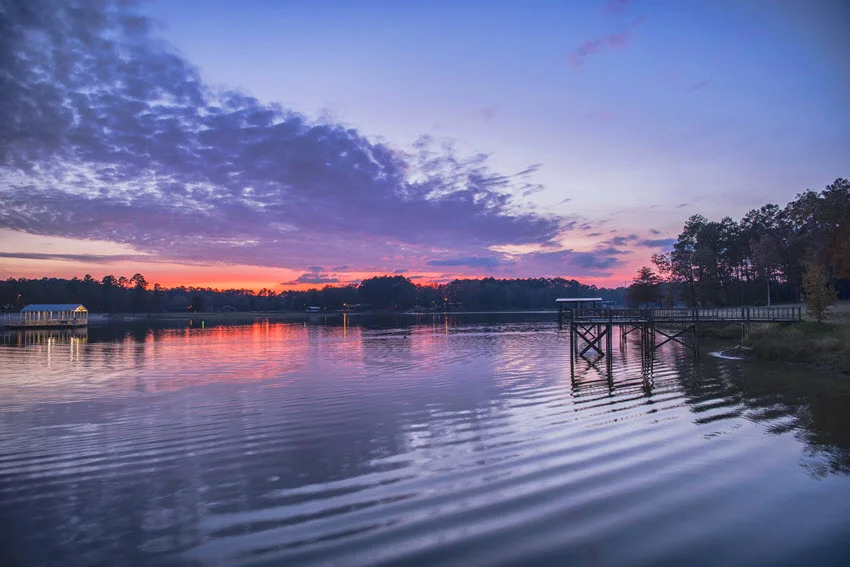 Pink and purple sundown over a pier at the Louisiana. The vibrant sky reflects on the calm water, casting colorful hues across the scene, while the silhouette of the pier stretches into the glowing horizon, creating a peaceful and dreamy atmosphere.