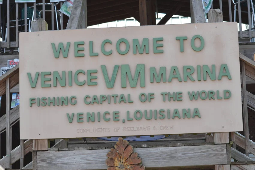 Street sign reading Welcome to Venice Marina in Venice, Louisiana, near New Orleans. The sign stands prominently against a backdrop of blue skies and marshland, with fishing boats and docks visible in the distance, capturing the vibrant atmosphere of this Gulf Coast fishing hub.