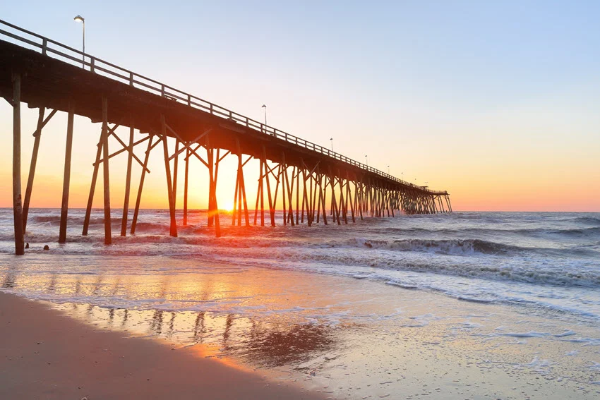 A venerable wood fishing pier at sunrise, stretching over calm waters. The soft, golden light of dawn bathes the pier, creating a serene and timeless atmosphere, perfect for early morning fishing or quiet reflection.
