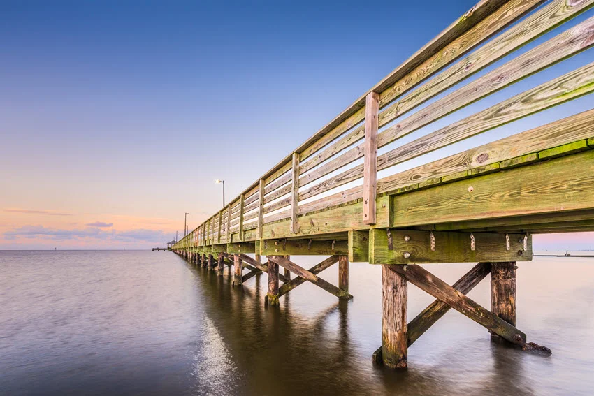 Lighthouse Pier in Biloxi, Mississippi, extending over the calm waters of the Gulf. The pier leads toward the horizon, with the iconic Biloxi Lighthouse visible nearby, set against a coastal backdrop of blue skies and gentle waves.