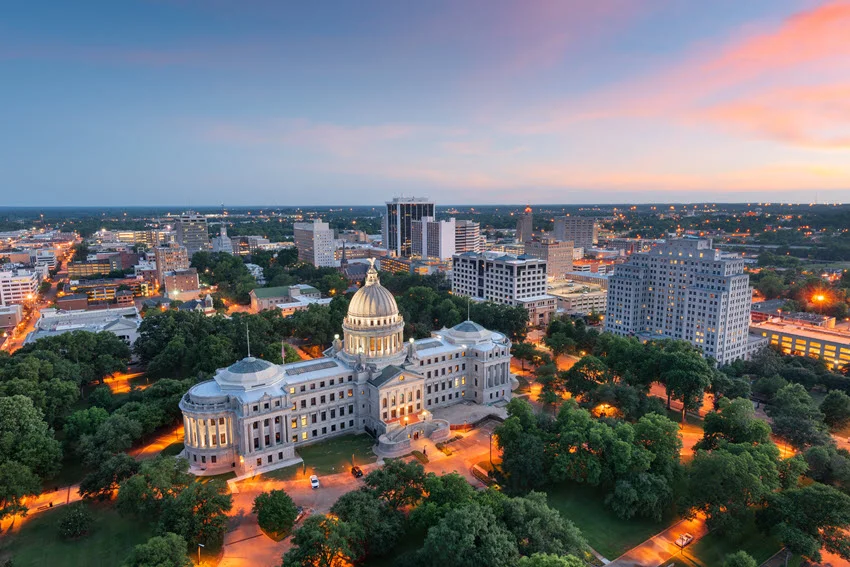 Jackson, Mississippi, USA skyline at dusk, featuring the illuminated Capitol Building. The cityscape glows with warm lights against the evening sky, highlighting the blend of historic and modern architecture.