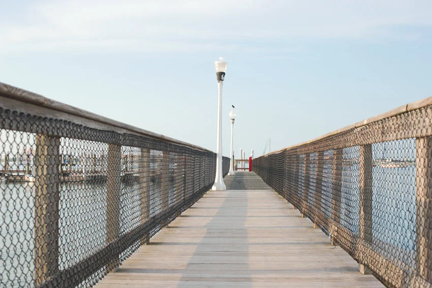 Low-angle view of a wooden boat dock, with leading lines guiding toward the water. A lamp post stands along the dock under a clear blue sky, with the early morning sun casting soft light on the scene.