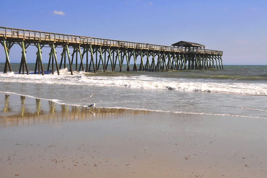 View from the beach of a long pier extending over gentle waves on a sandy beach near Ocean Springs, Mississippi.