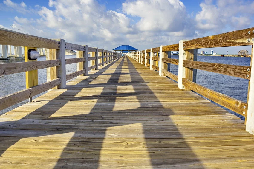 A sunny view of a wide wooden pier with railings leading out over the water in Ocean Springs. The wooden pier stretches over calm, reflective waters with clear skies above, creating a peaceful and scenic coastal view.