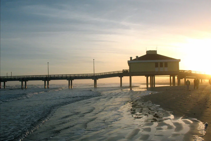Backlit pier at Aransas Pass, Texas, with the sun casting a golden glow behind the wooden structure. The light filters through the pier's beams, creating a stunning contrast against the calm coastal waters and sky.