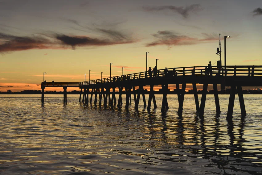 Fishing pier in downtown Sarasota at sunset, with city skyline silhouettes and golden reflections over the calm bay creating a peaceful coastal view.