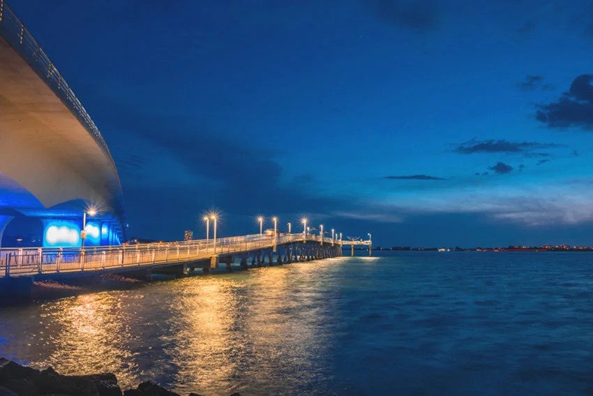 Fishing pier in Sarasota Bay, Florida, with the Ringling Bridge in the background under a dramatic evening sky and calm coastal waters.