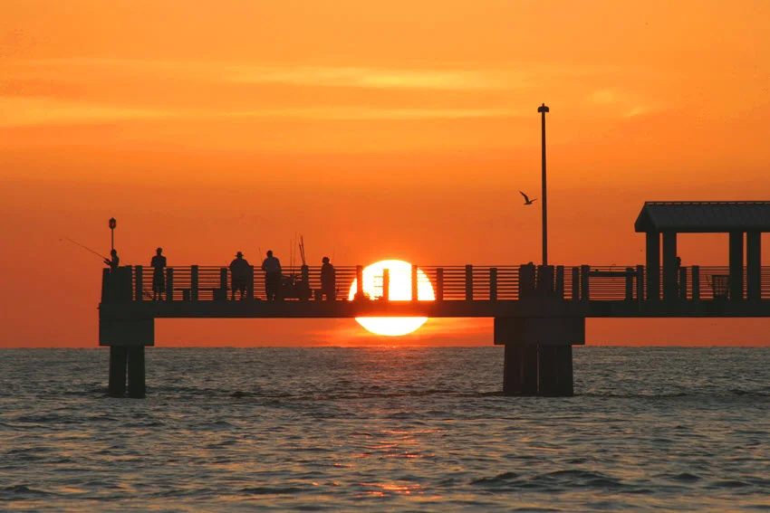 ibrant Gulf Coast sunset over Fort De Soto Pier in Saint Petersburg, Florida. The sky is painted with shades of orange, pink, and purple, creating a stunning backdrop for the silhouette of the pier, perfectly reflecting the serene beauty of the coastline.