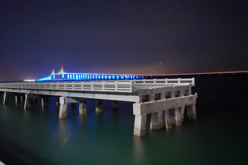 Night landscape of Skyway Fishing Pier State Park in Florida, showcasing the old, collapsed bridge that has been converted into the longest fishing pier in the world. The pier stretches into the dark waters, illuminated by lights that highlight the dramatic structure, creating a unique and tranquil fishing experience.
