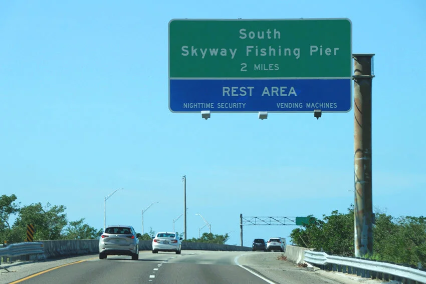 View of the exit leading to the South Skyway Fishing Pier and Rest Area in St. Petersburg, Florida. The pathway is surrounded by lush greenery, guiding visitors toward a well-known fishing and relaxation destination.