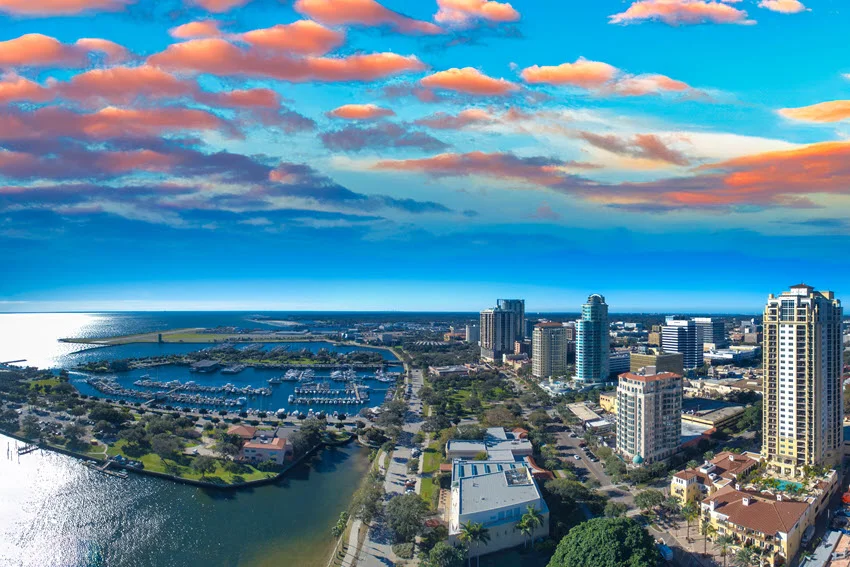 Panoramic aerial view of the St. Petersburg skyline at sunset in Florida. The warm glow of the setting sun lights up the cityscape, with Tampa Bay's shimmering waters enhancing the vibrant charm of this coastal destination.