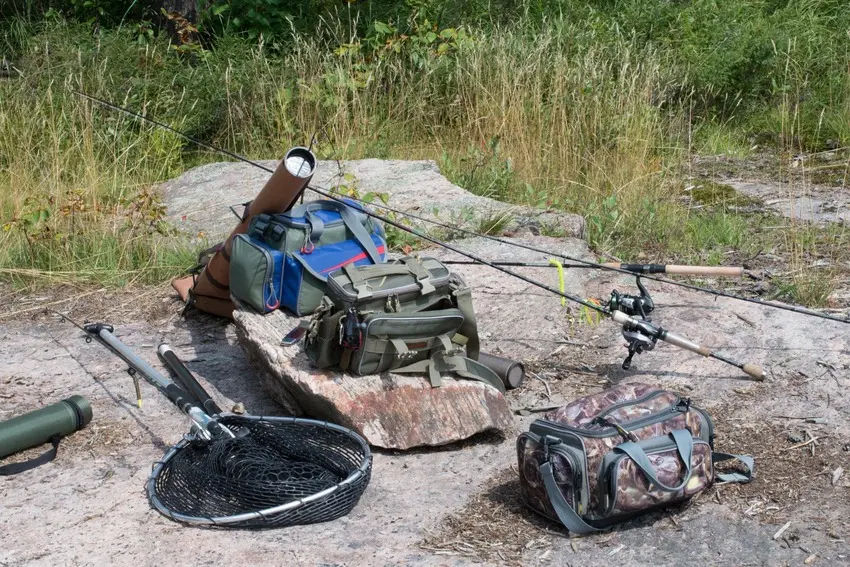 Fishing bags, rods, and landing nets arranged on rocks near a grassy lakeshore.