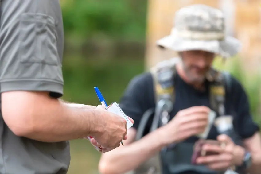 A ranger checks an angler's fishing license during a routine inspection beside a lake, with fishing gear visible nearby.
