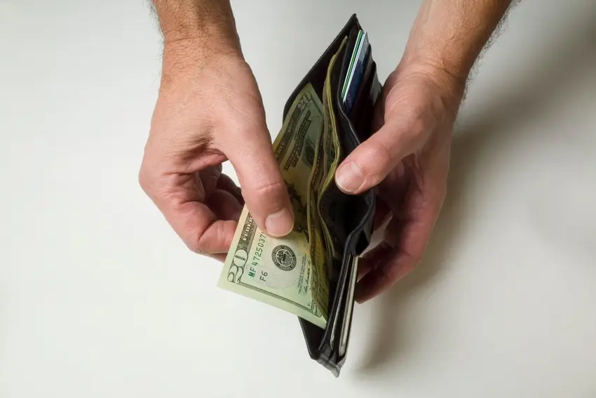 Hands remove US dollar bills from a wallet against a clean white background, with several banknotes visible inside.