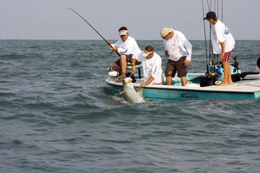 Anglers on a fishing boat struggle with a Tarpon caught on the hook, battling the fish in the open water.