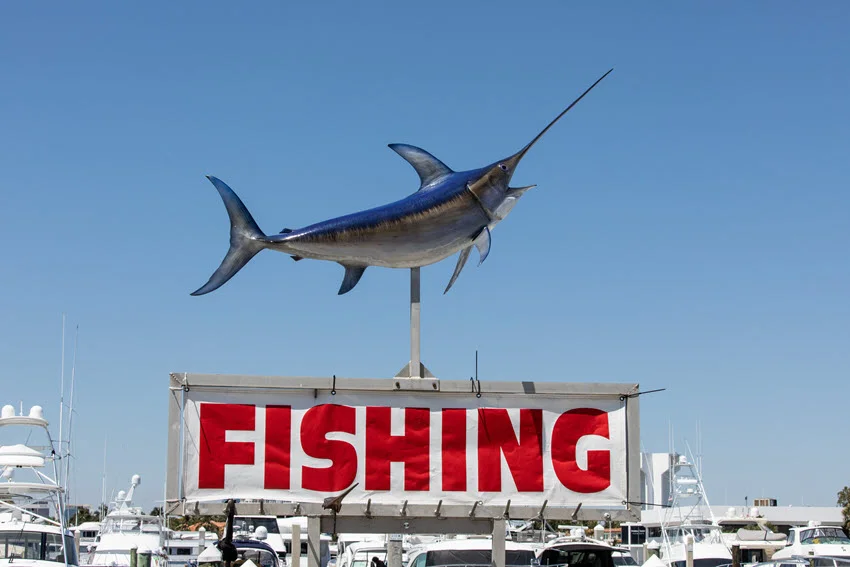 A model spearfish mounted on a vibrant sign advertising deep-sea fishing, located at a bustling pier in Fort Lauderdale.