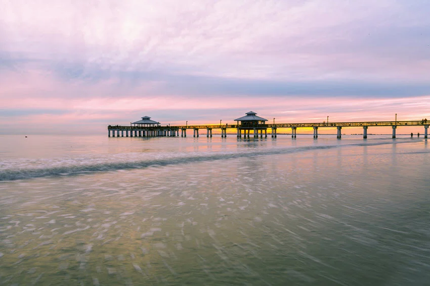 A distant view of the fishing pier at Fort Myers Beach, illuminated by warm evening light, with the calm waters of the Gulf of Mexico and a serene Florida sky.