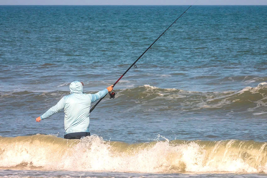 A lone coastal fisherman with a strong grip keeps his balance while a wave breaks behind him along a beach on a sunny afternoon in St. Augustine.