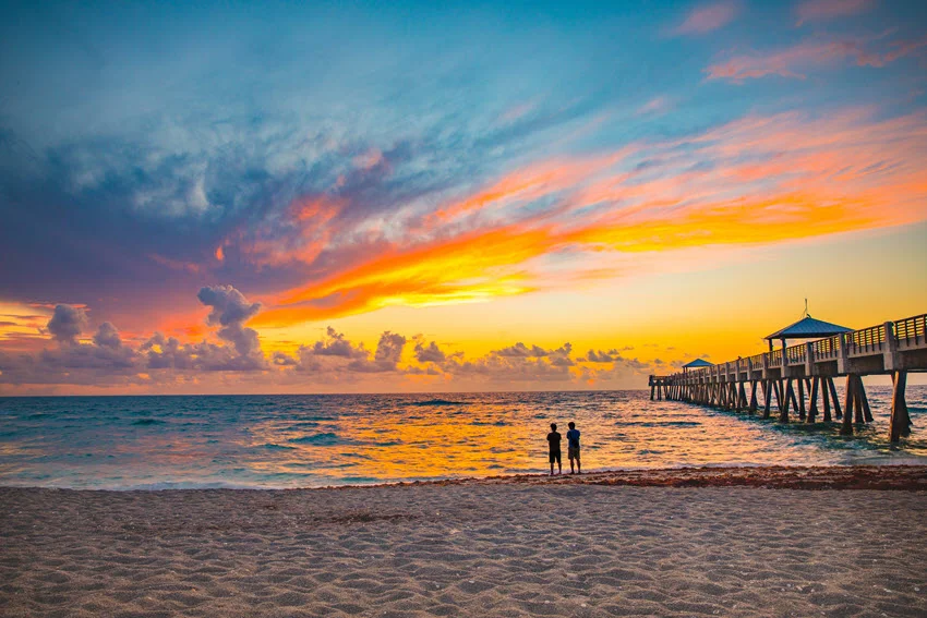 Two people standing on the beach next to the fishing pier watching the beautiful sunrise over the ocean.