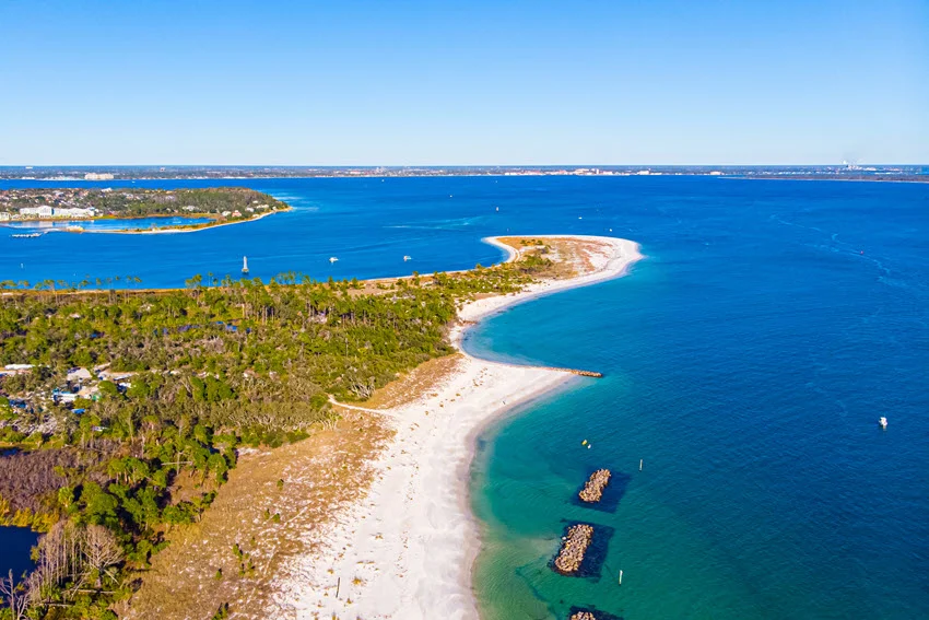 Aerial shot of the beach and curved coastline at St. Andrews State Park in Florida, showing clear blue water, sandy shoreline, and a scenic coastal fishing area.